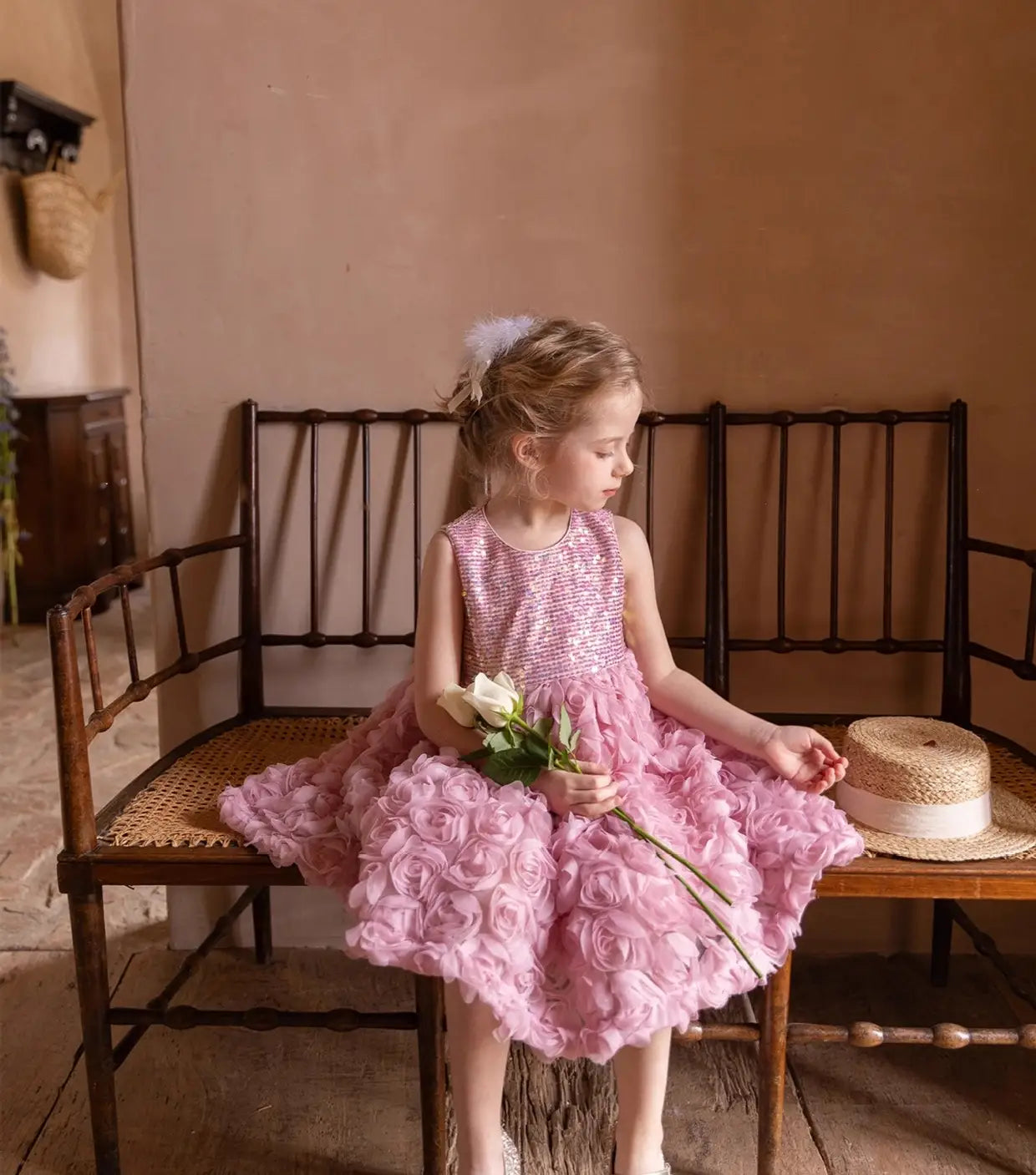 Young girl in a pink dress sitting on a wooden bench holding flowers.
