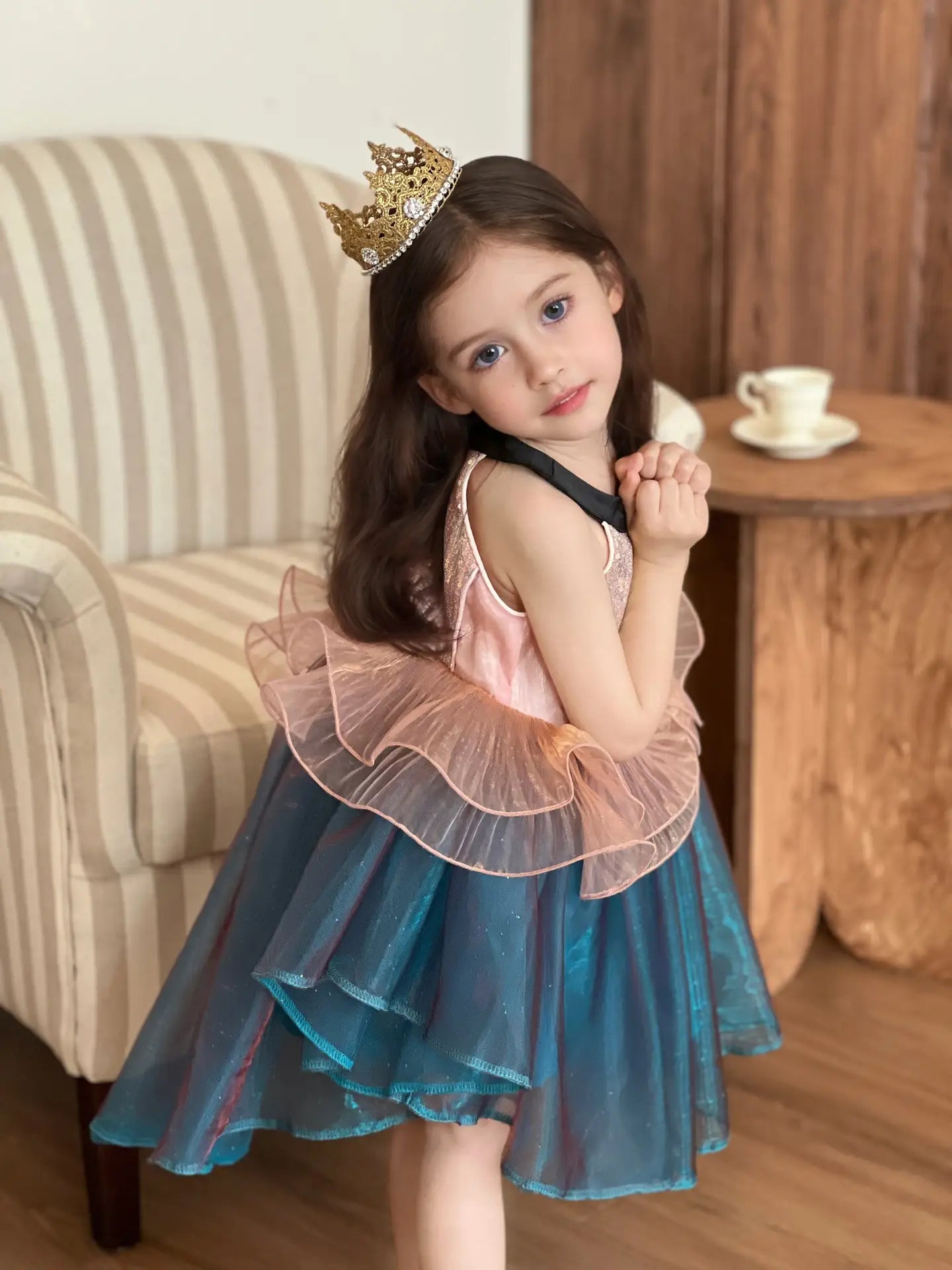 Young girl in a blue and pink Sleeveless Sea Princess Sequin Blue Tulle  Mermaid Dress with a crown, sitting on a striped chair.