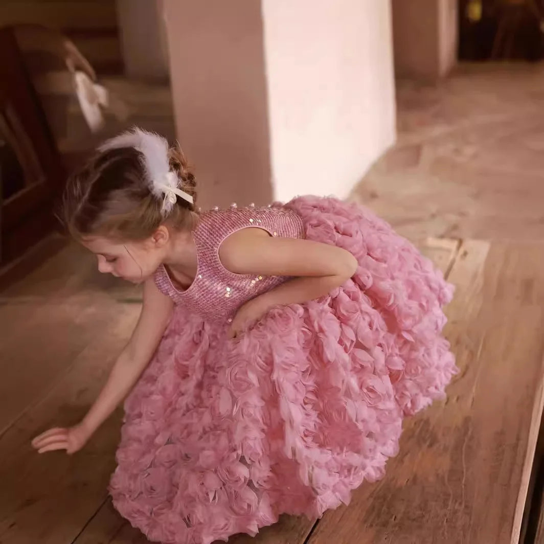 Young girl in a pink dress with floral details and sequins, standing on a wooden floor.