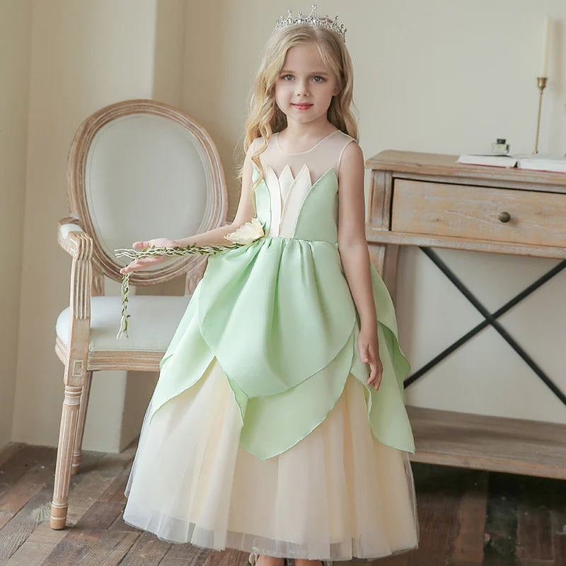 Young girl in a green and beige Green Lily Flower frog Princess Dress with a tiara, standing in a room with a chair and wooden desk.