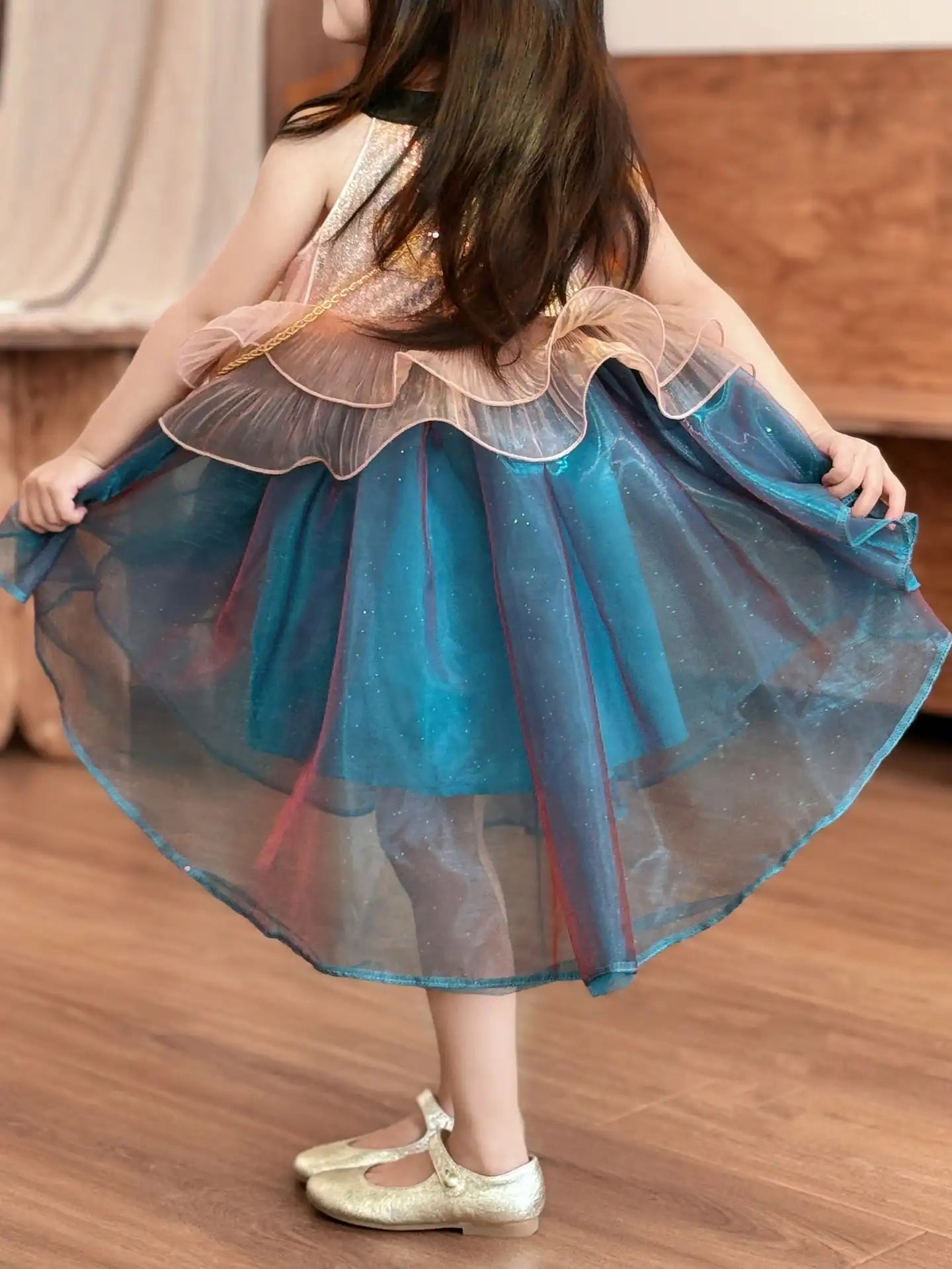Child wearing a blue and beige dress with a ruffled top on a wooden floor.