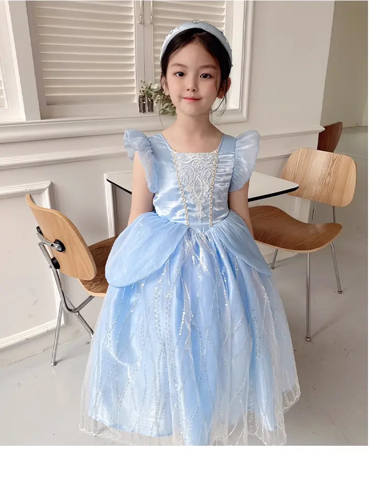 Young girl in a light Blue Fairy Princess Dress for Girls , Elegant Tulle Party Gown standing in a room with wooden chairs and a white wall.