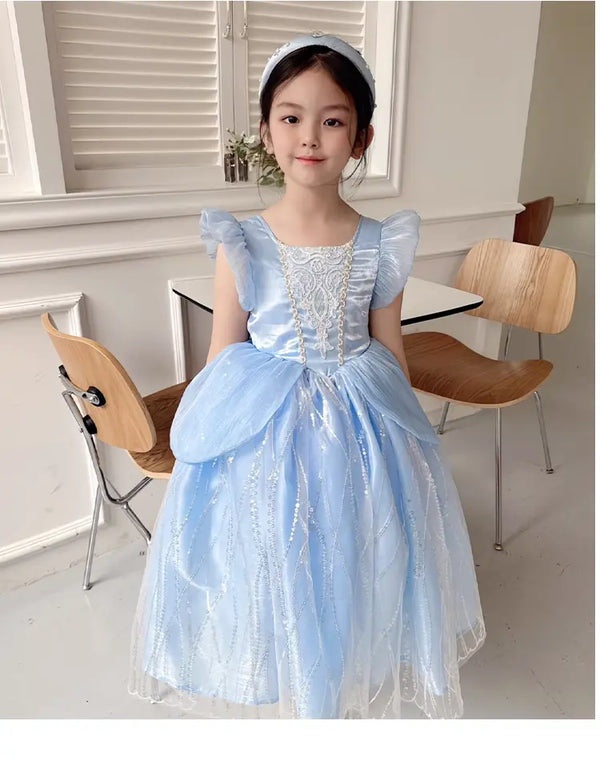 Young girl in a light Blue Fairy Princess Dress for Girls , Elegant Tulle Party Gown standing in a room with wooden chairs and a white wall.