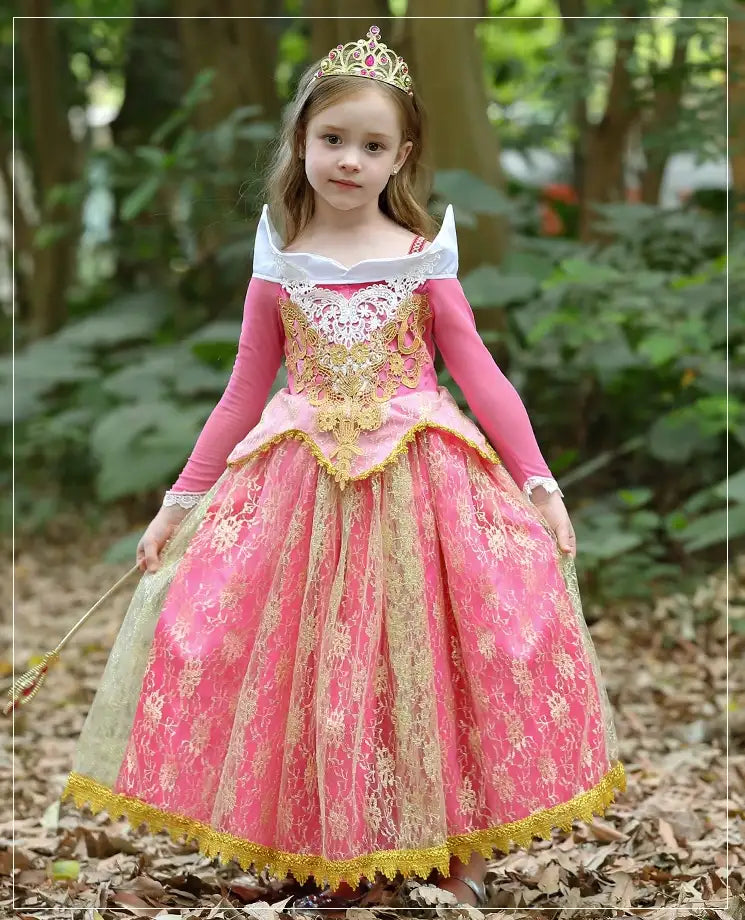 Young girl in a pink and gold princess dress with a tiara, standing outdoors among trees.