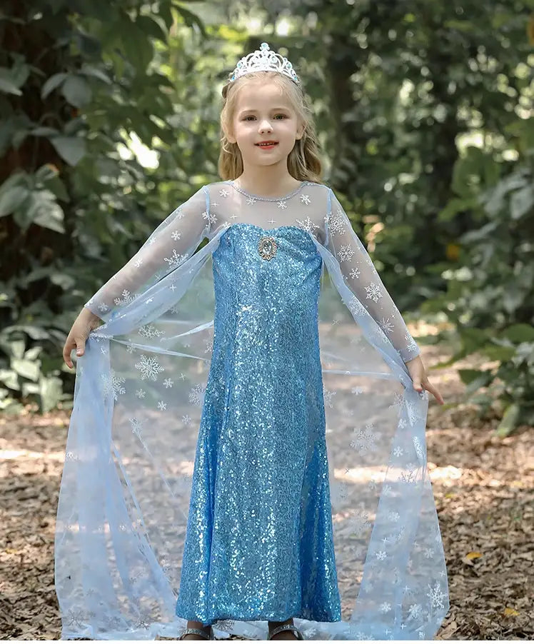 Young girl in a  Glittering Winter Queen Blue Sequin with Snowflake Cape dress with a tiara standing outdoors among trees.