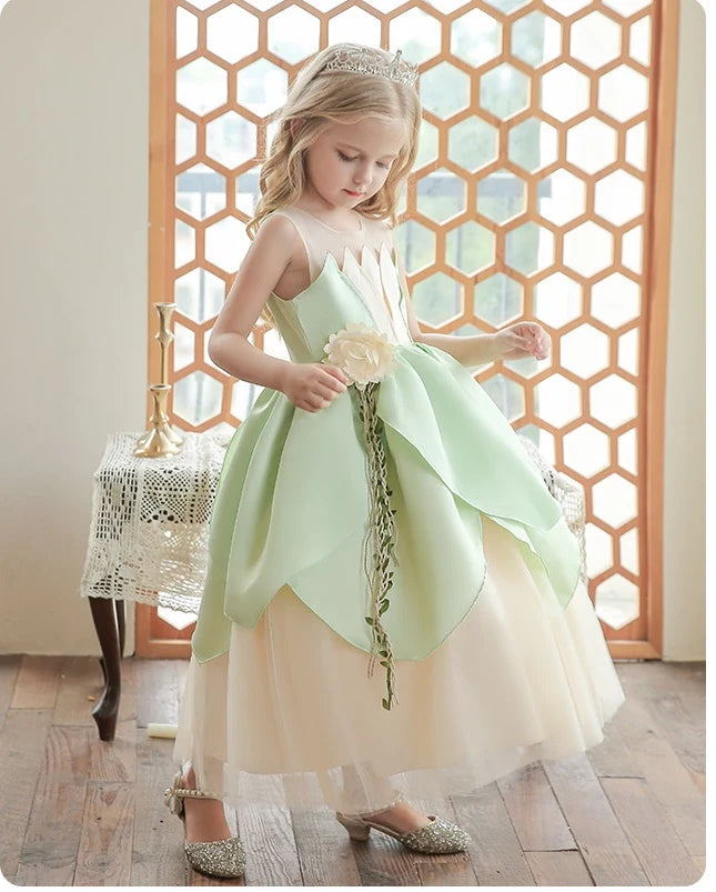 Young girl in a light green and beige  Lily Flower Princess Dress holding a flower, standing in front of a decorative screen.