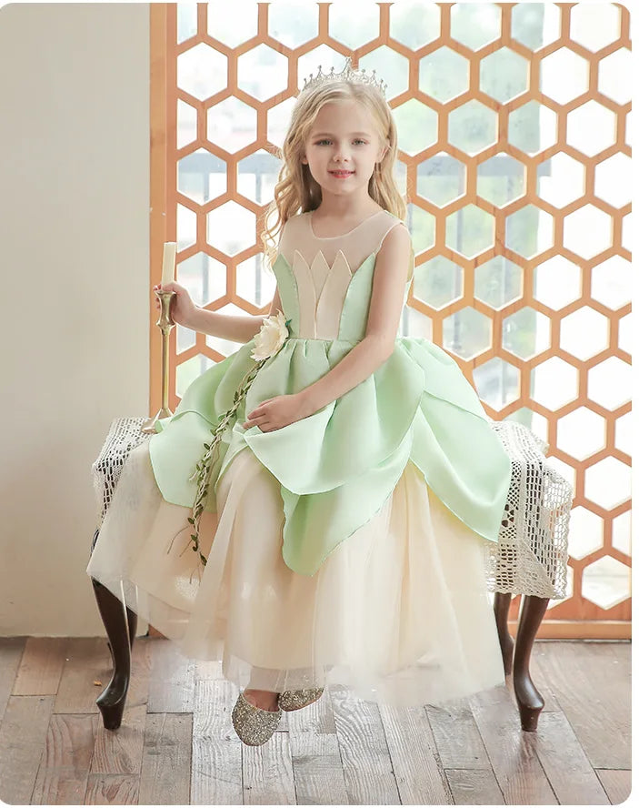 Young girl in a light green and beige frog princess dress sitting on a decorative stool with a geometric wall in the background.