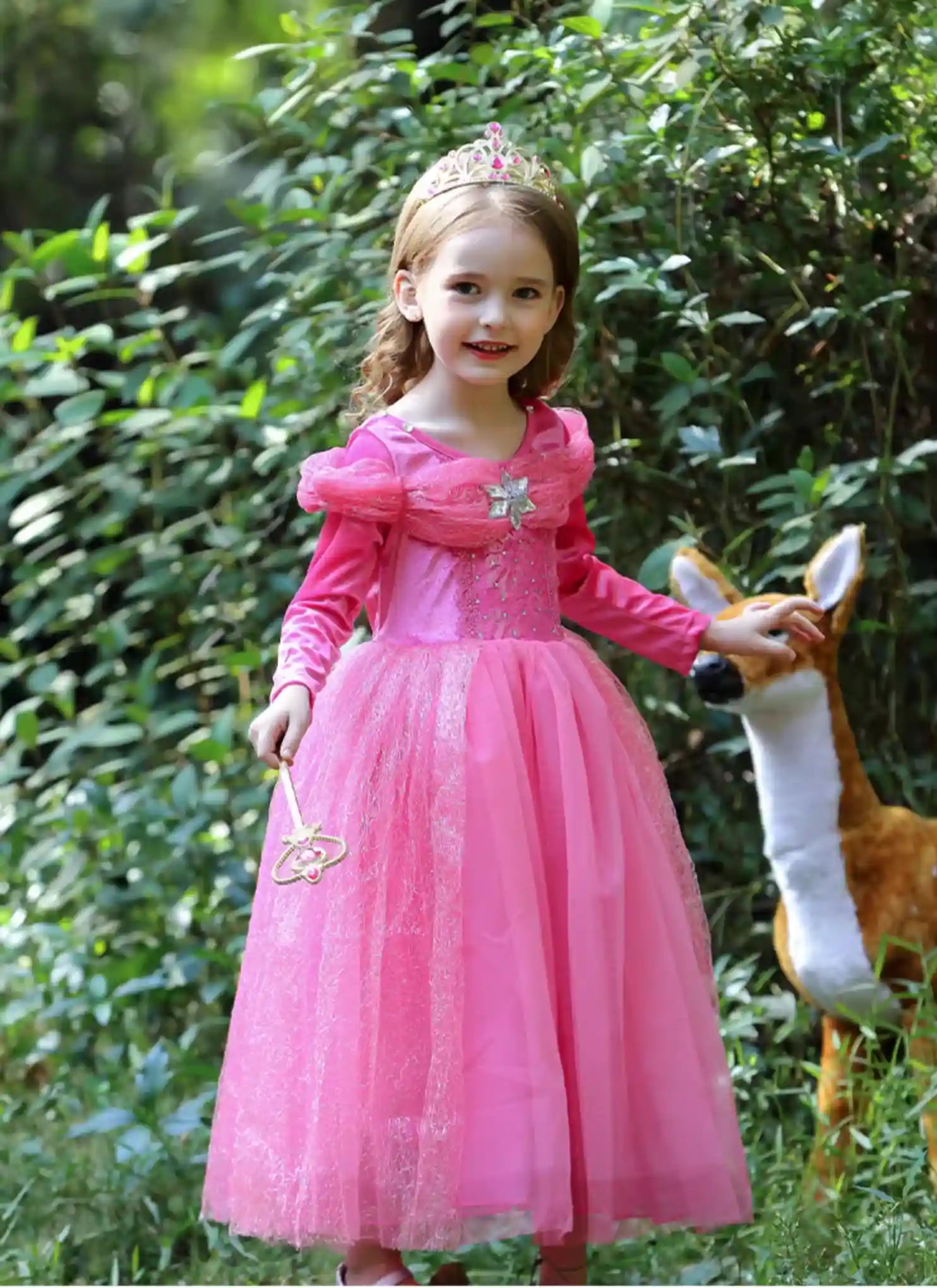 Young girl in a pink princess dress with a tiara standing outdoors with a deer.