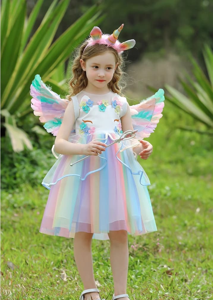 Child in a Rainbow Unicorn Fairy Dress costume with wings and headband standing in a grassy area.