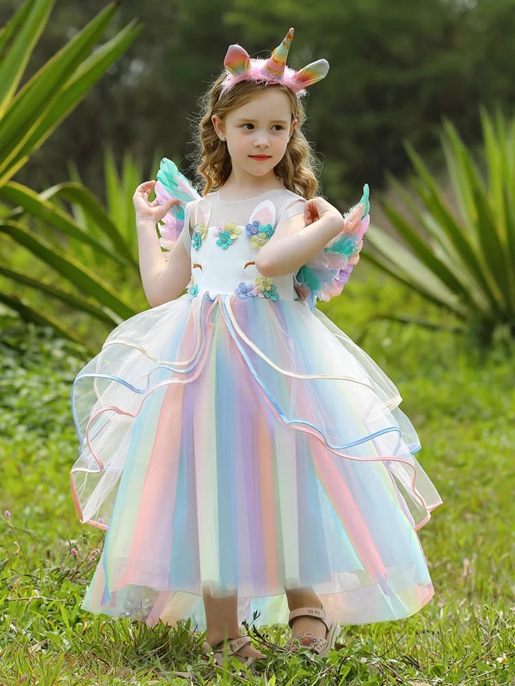 Young girl in a unicorn-themed dress with wings and headband standing in grass.