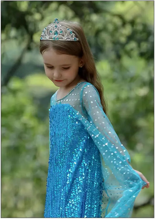 Young girl in a Sparkling Ice Queen Blue Sequin Gown for Girls with Sheer Cape  with a tiara, standing outdoors with greenery in the background