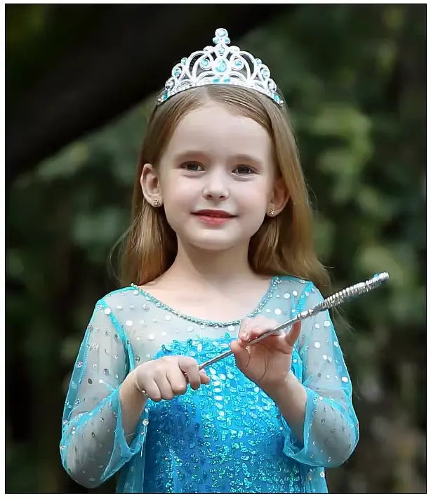 Young girl in a Sparkling Ice Queen Blue Sequin Gown for Girls with Sheer Cape 4with a tiara and wand, standing outdoors.