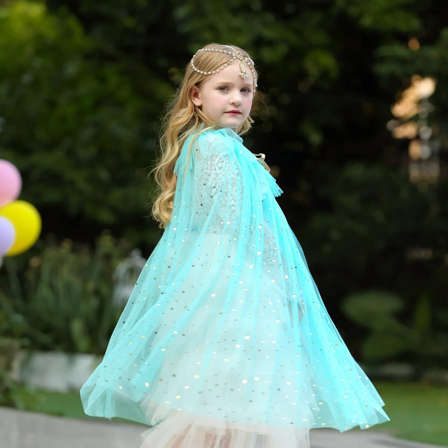 Young girl in a turquoise Star Tulle Fairy Dress Up Princess Cape  and headband, standing outdoors with balloons in the background.