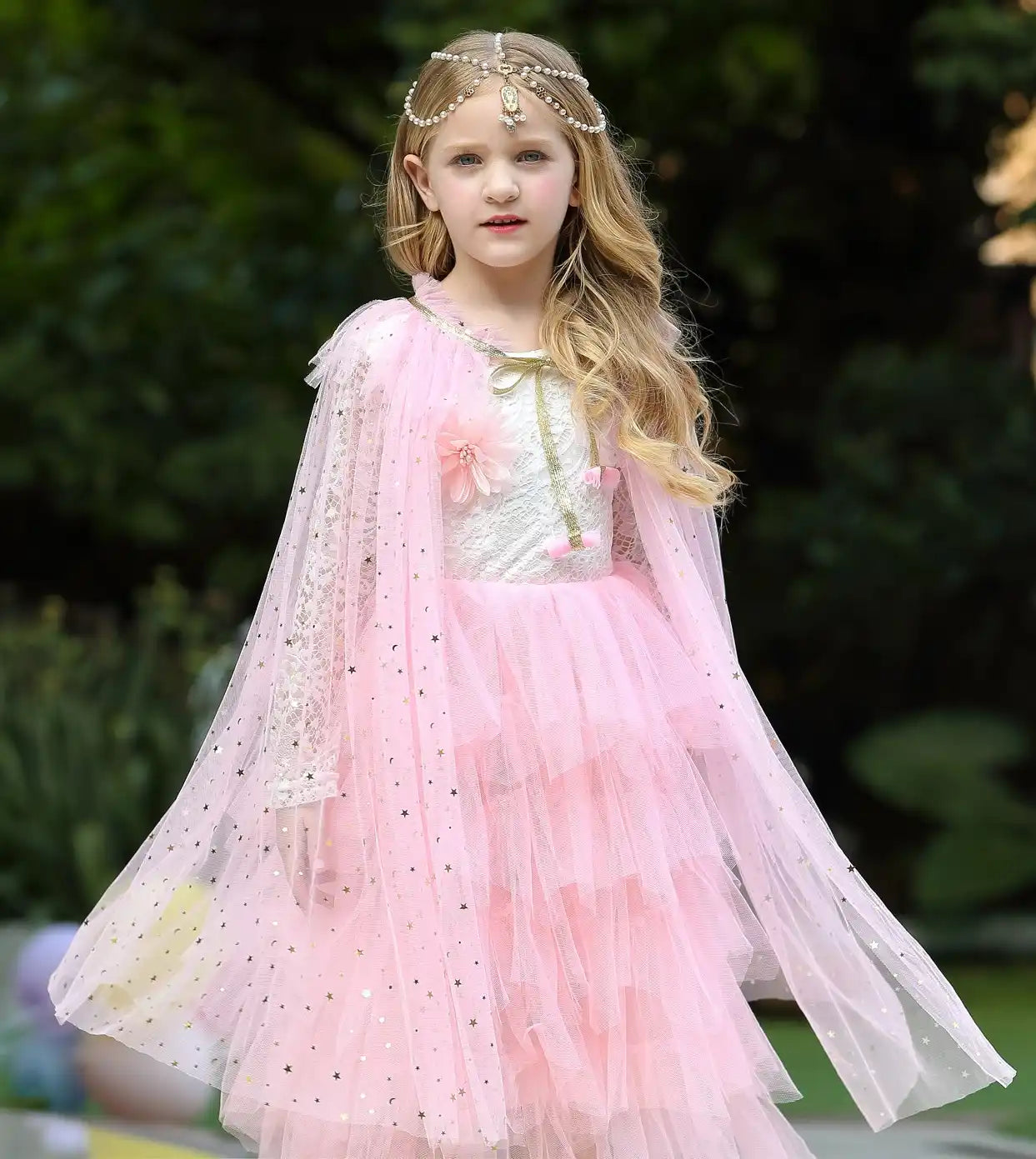 Young girl in a pink dress with a floral top and pink Star Tulle Fairy Dress Up Princess Cape, standing outdoors.