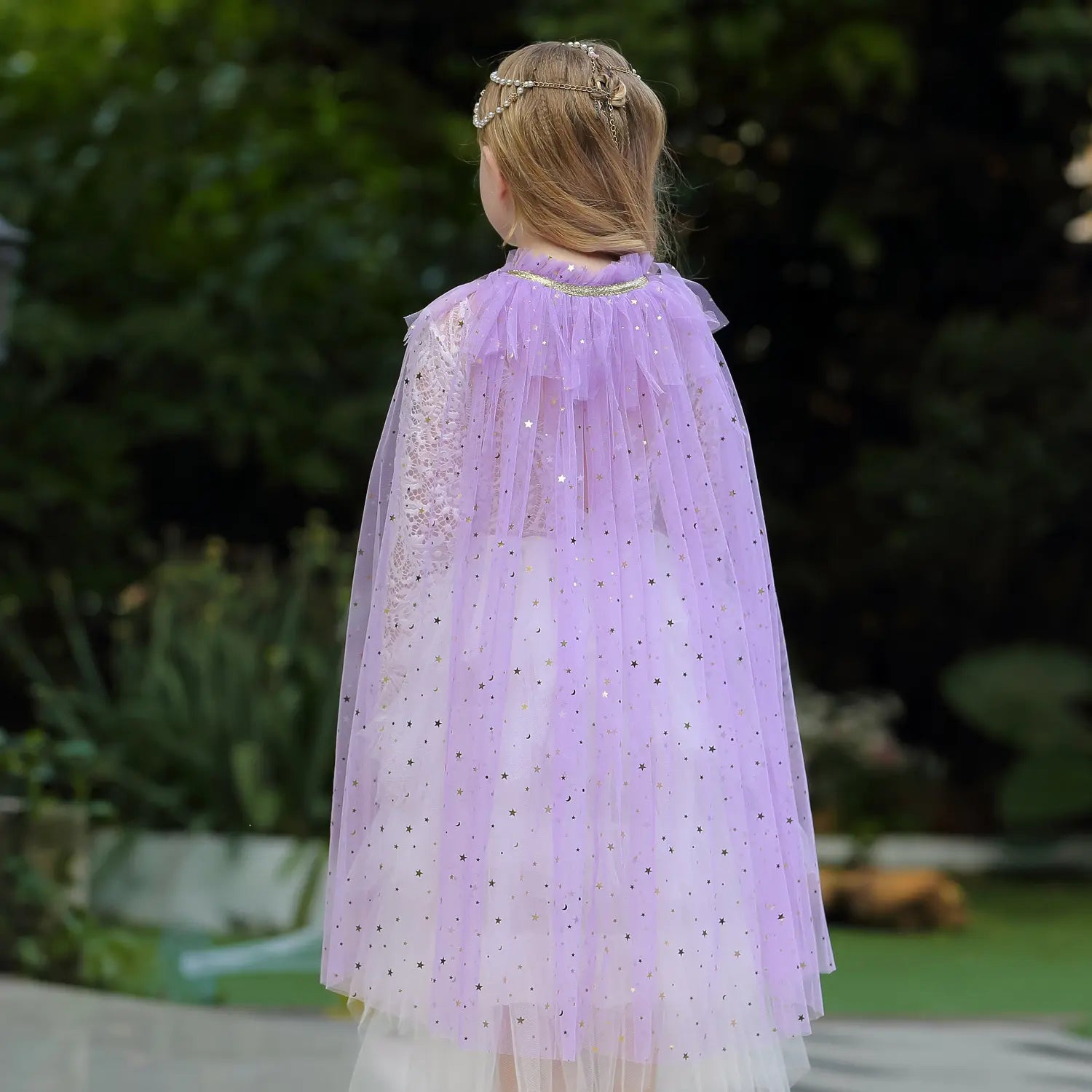 Child wearing a purple tulle cape outdoors with greenery in the background