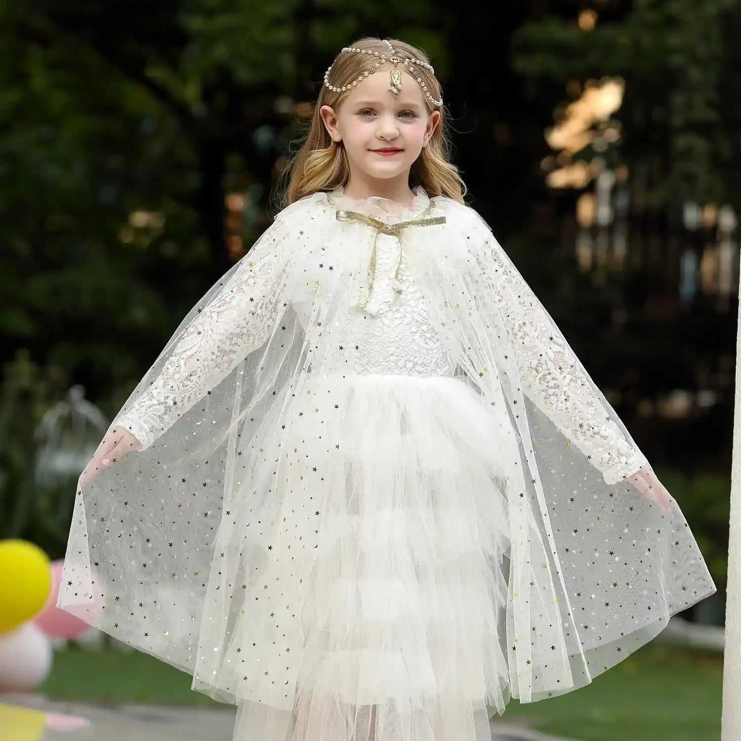 Young girl in a white Star Tulle Fairy Dress Up Princess Cape  and headband, standing outdoors.